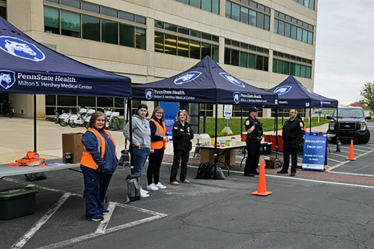 Six people stand near canopies with Penn State Health logos on them. A large building is in the background.