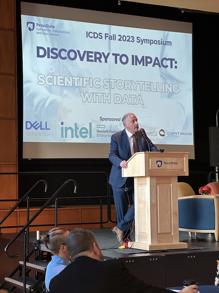 Andrew Read, Senior Vice President for Research, stands on stage at a wooden podium presenting opening remarks.