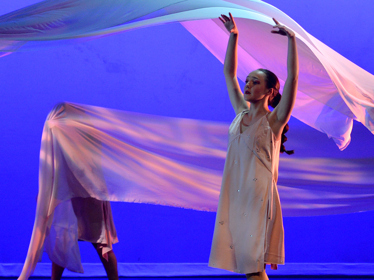 A dancer in the foreground with her arms raised in the air while a piece of fabric flows in the air behind her