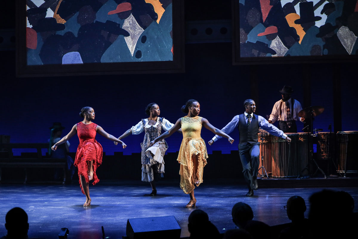 Black dancers strike matching poses while a backdrop shows bold paintings.