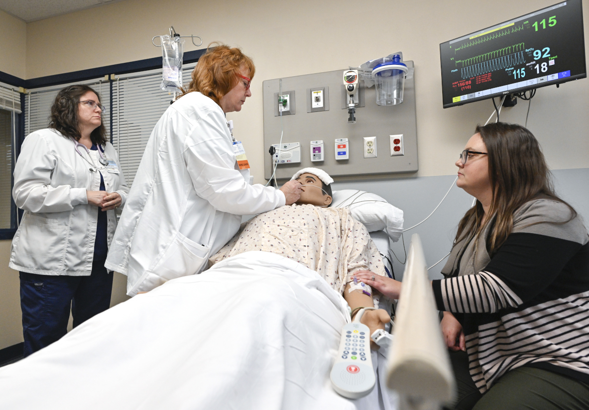 Two nurses tend to a mannequin posing as a patient while a woman sits at the bedside, looking on.