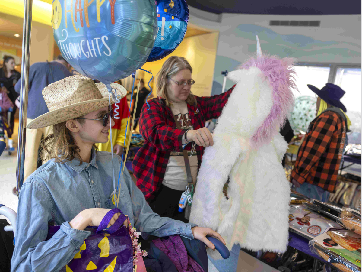 A mother and her teenage son peruse costumes and other items laying on tables. The son, seated in a wheelchair, holds balloons and other items.