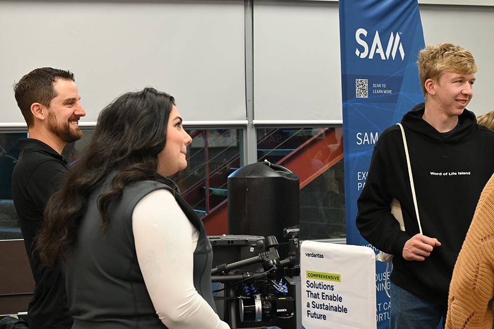 Adults and students conversing during a career fair, with a flag with the SAM logo at back right.