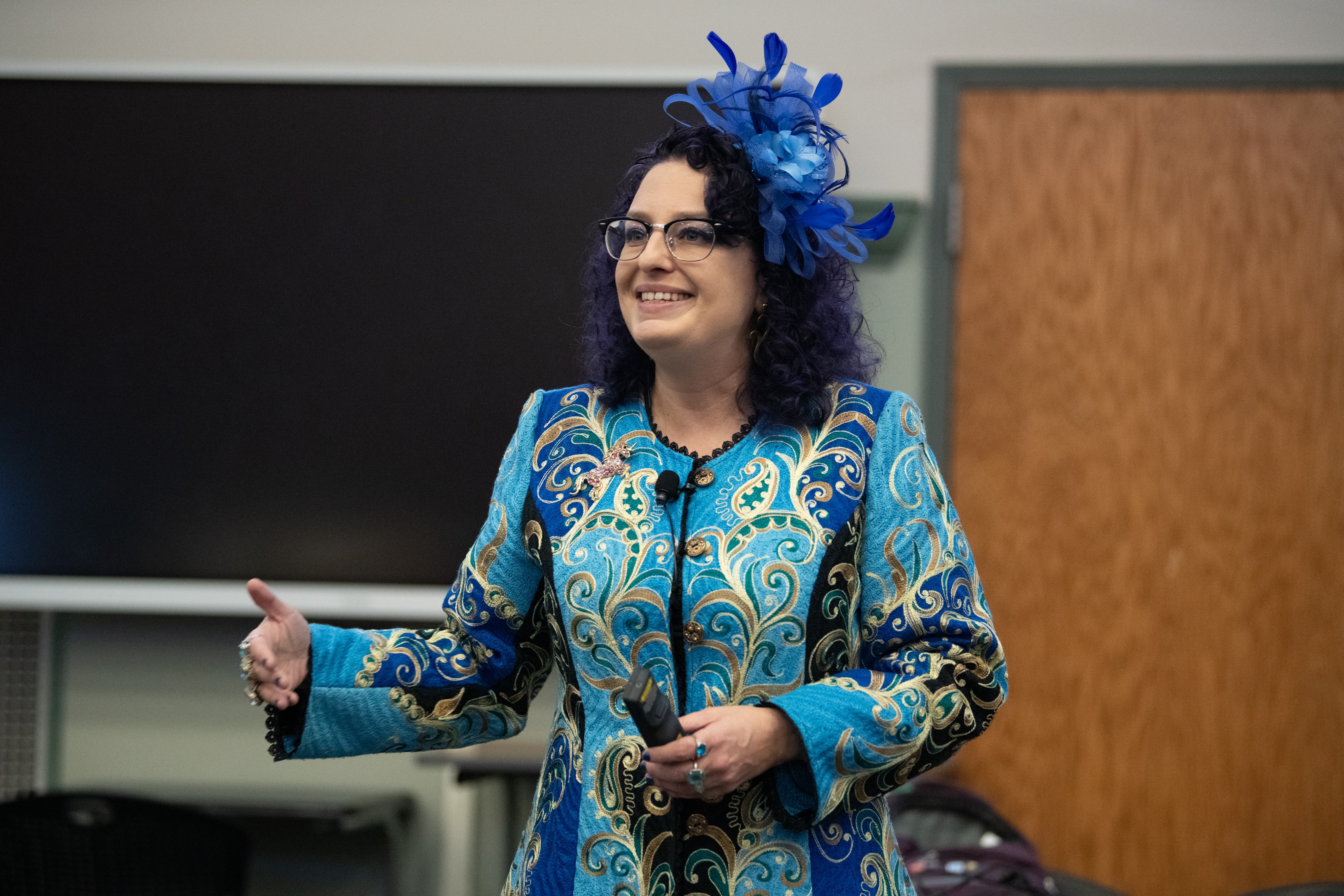 A person wearing a blue fascinator stands in front of a blackboard and a door.