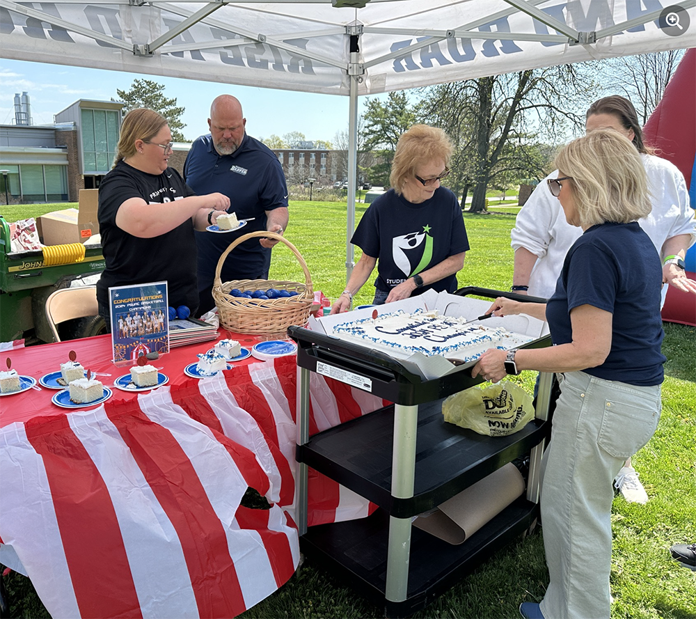 A group of people stand at a table and prepare to serve pieces of cake under a tent