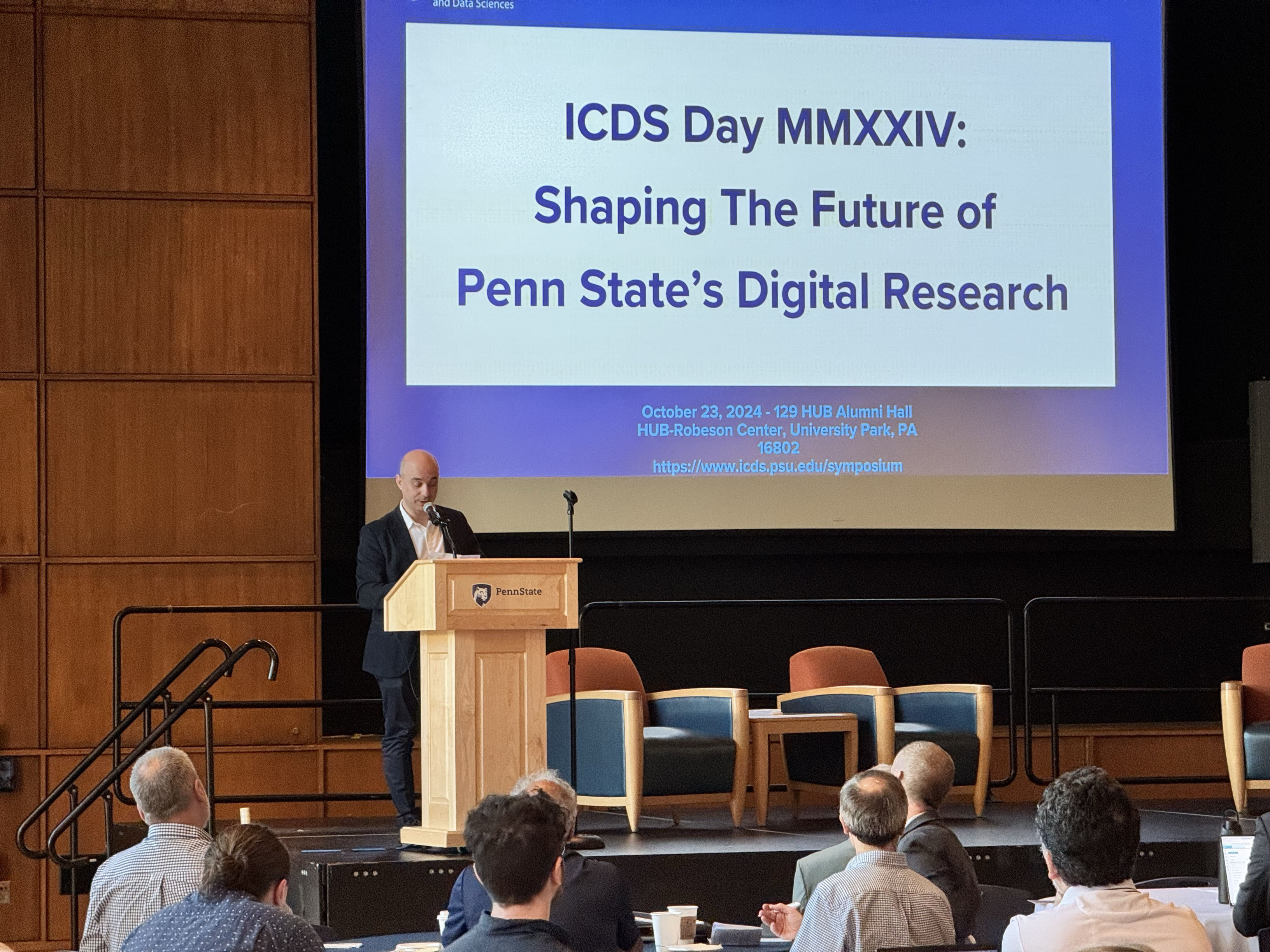 Guido Cervone, Penn State Institute for Computational and Data Sciences (ICDS) stands at a wooden podium on the left as he presents a welcoming speech on stage at the HUB-Robeson Center.