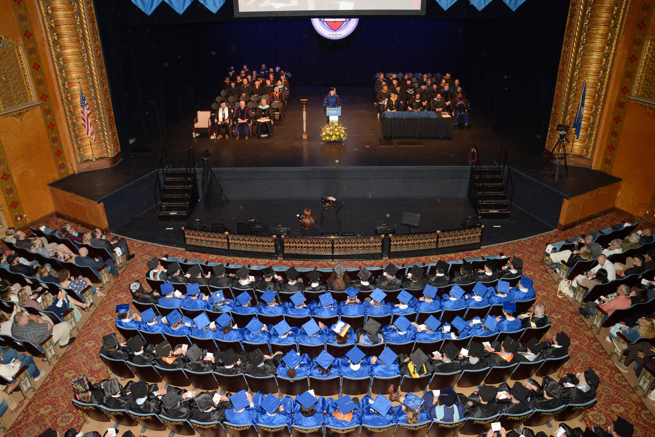 A balcony view of a theater stage and seats filled with people in blue graduation caps and gowns