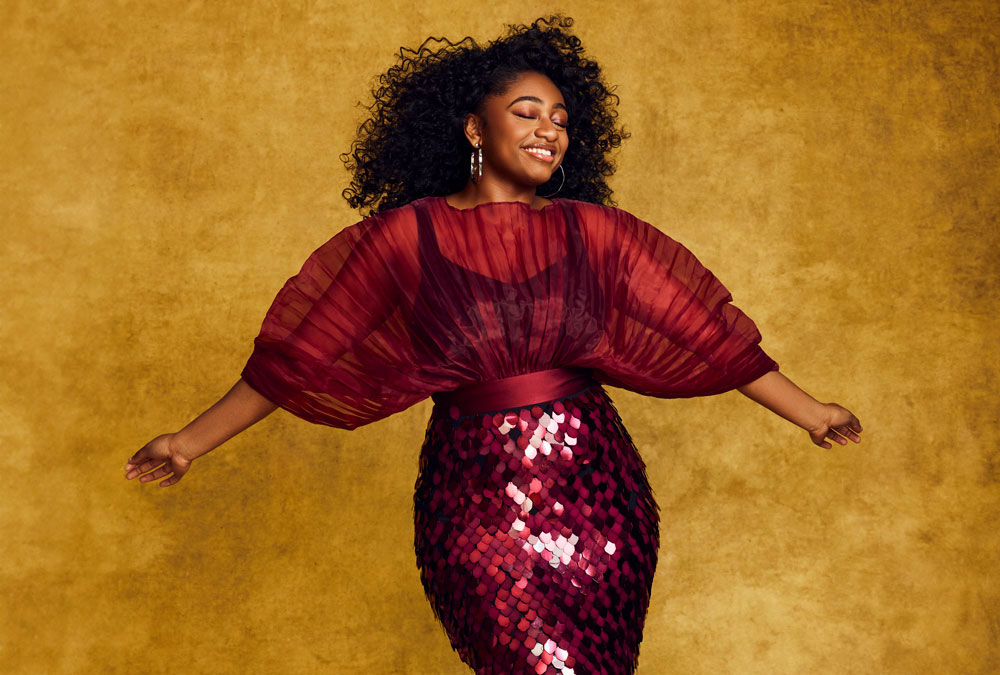 A young Black woman wearing a sequinned dress stretches out her arms.