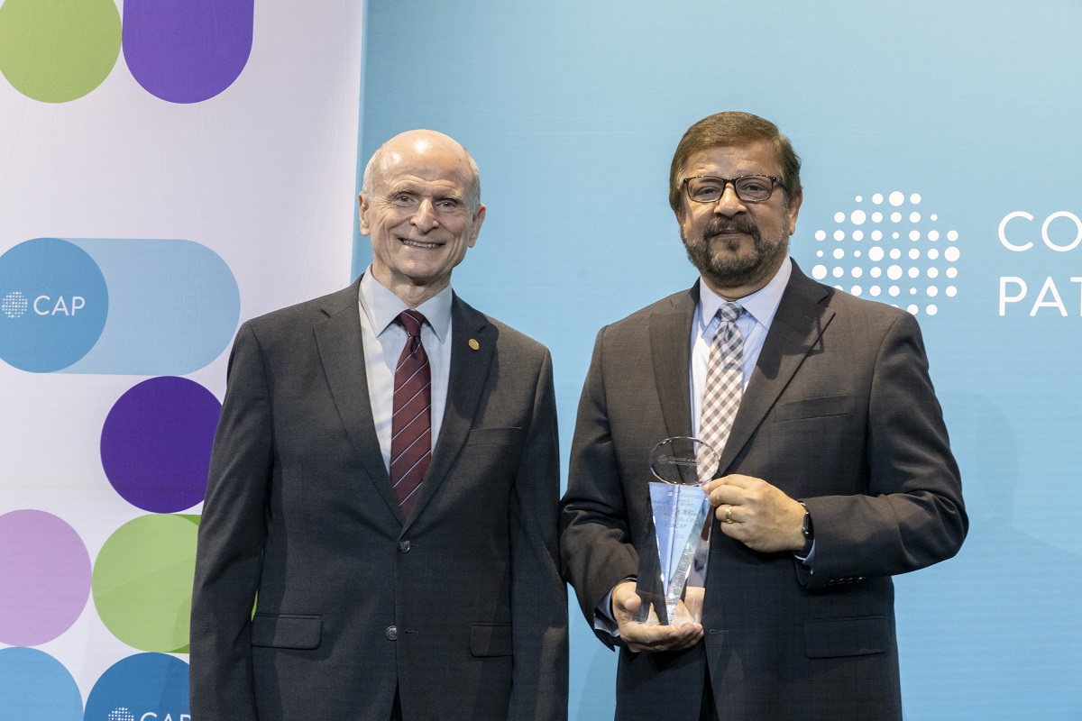 Two men stand on a stage posing for a photo. The man on the right is holding an award.