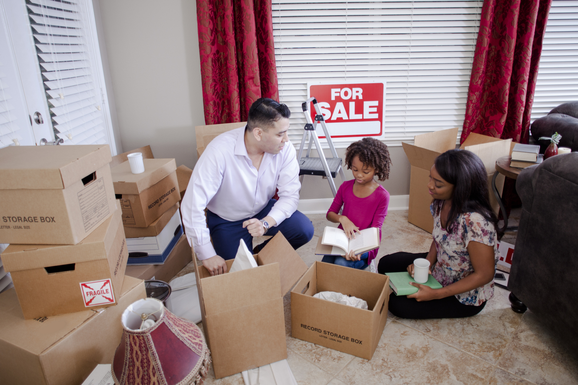 Multicultural family members pack boxes in new home. “For Sale” sign hangs on wall..