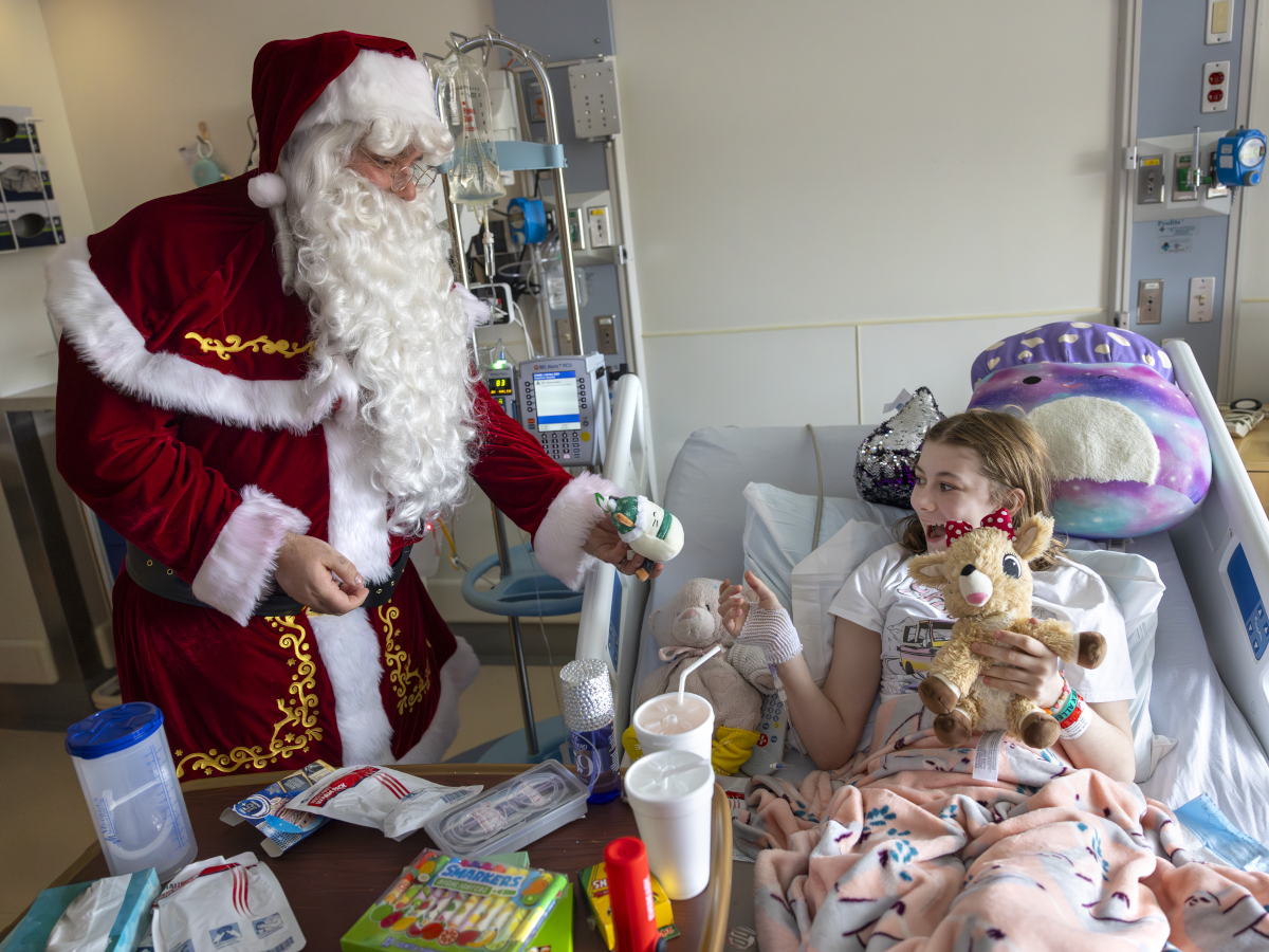 A young girl in a hospital bed, in a room filled with medical equipment and other supplies, smiles as she reached out to accept a gift of a small stuffed animal from Santa Claus.