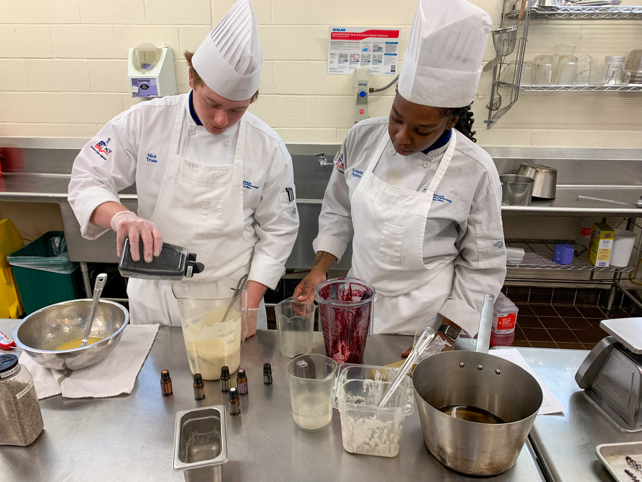 Two students in white kitchen uniforms and hats work over a pot on a stove