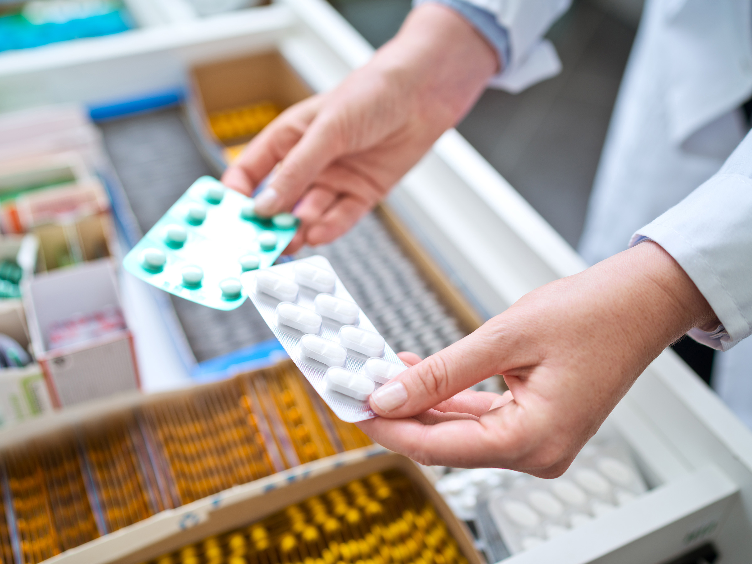 Two packs of pills being held above a drawer containing more pharmaceuticals 