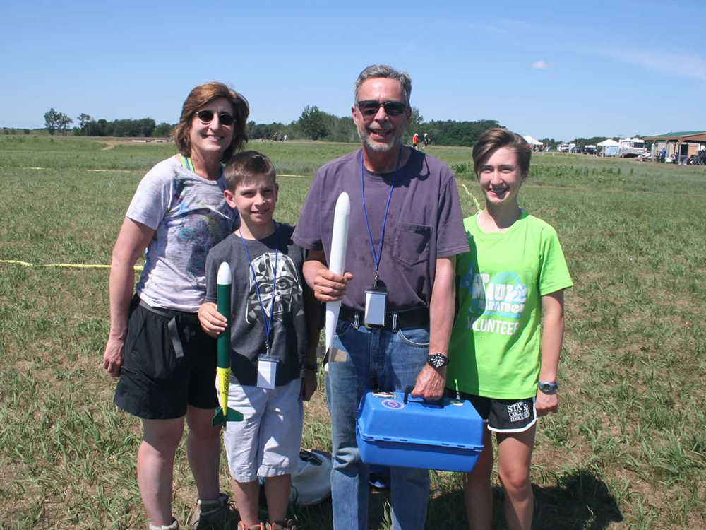 A family portrait of two adults and two kids standing in a field holding model rockets. 