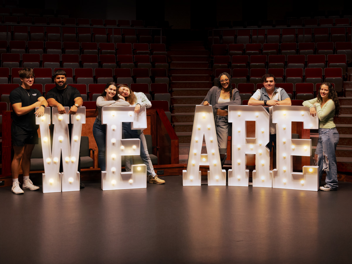 New Kensington students standing behind large letters that spell "WE ARE" inside the campus theatre.