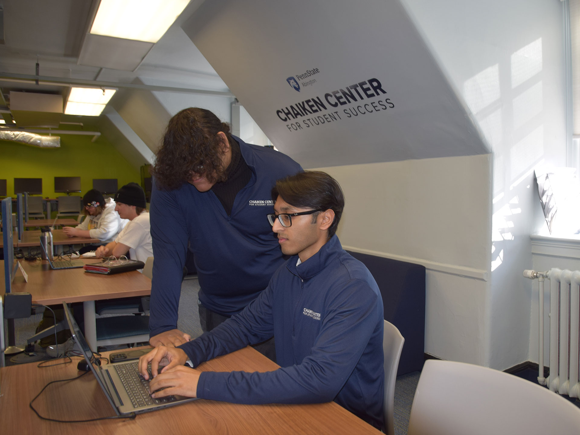 Person standing and person sitting and looking at a laptop