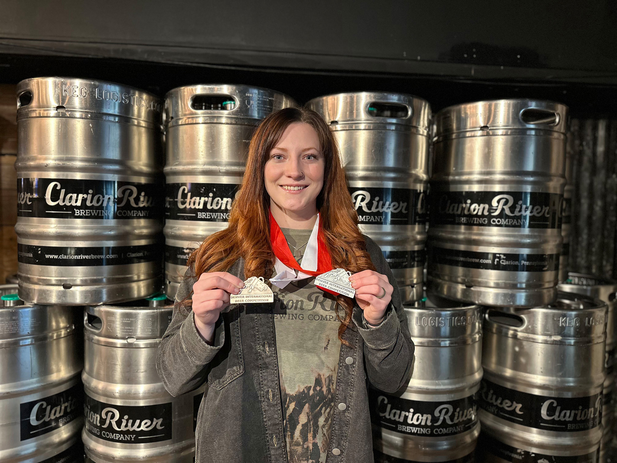 A person stands holding two medals in front of a wall of kegs at a brewing company