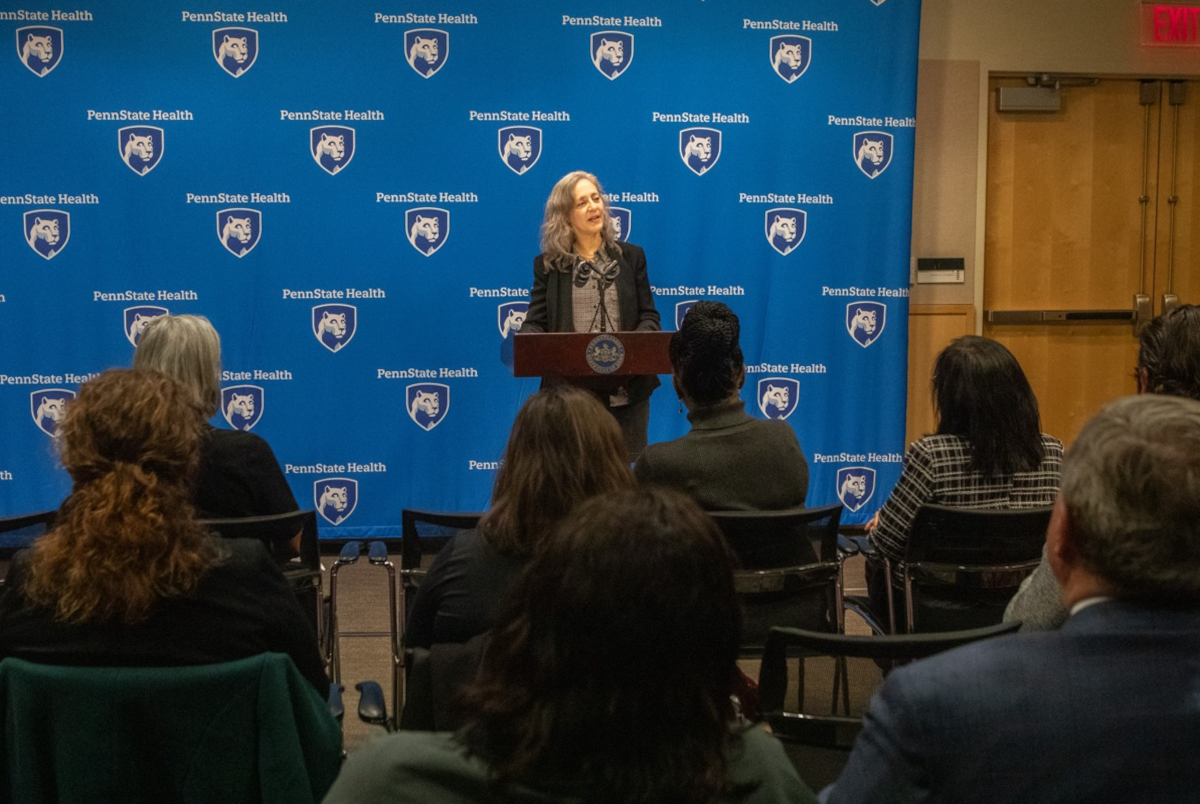 A woman in professional attire speaks at a lectern, gesturing with her hands. A step-and-repeat ‘Penn State Health’ backdrop is in the background.