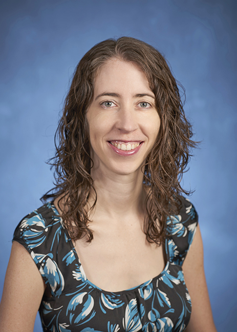 Erica Frankenberg head shot with long, wavy brown hair, black, blue, and green print short-sleeved blouse, smiling.