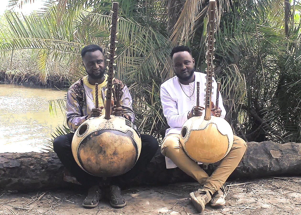 Two musicians pose outside near a river with their instruments