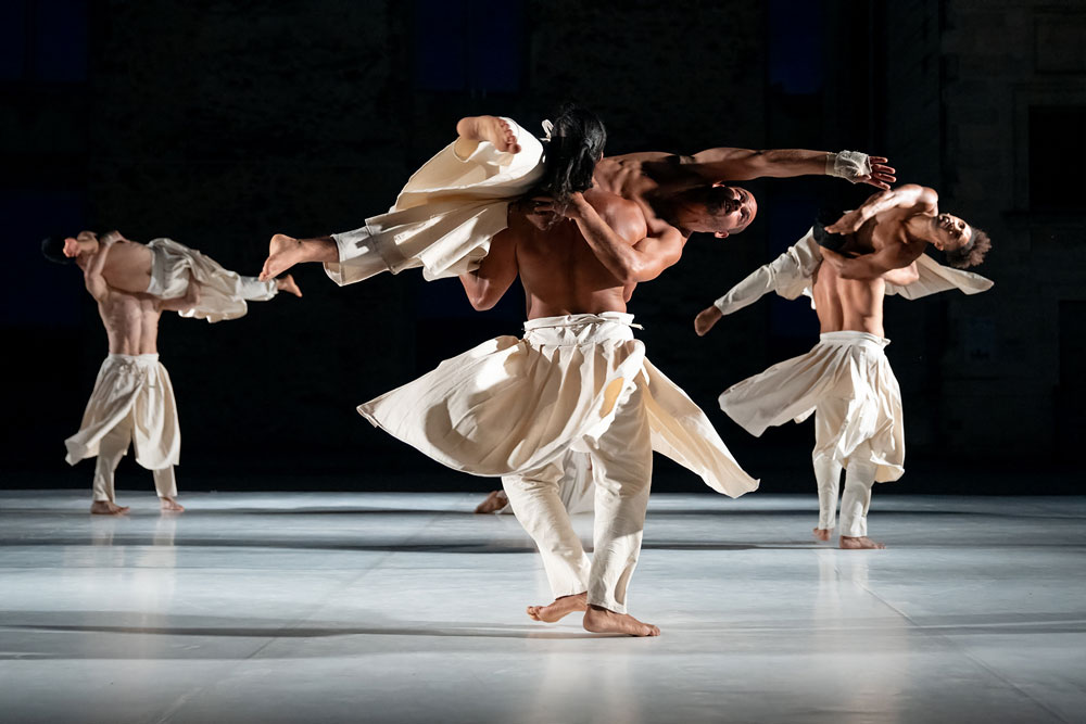 Three shirtless men wearing flowing robes each twirl another man on their shoulders.