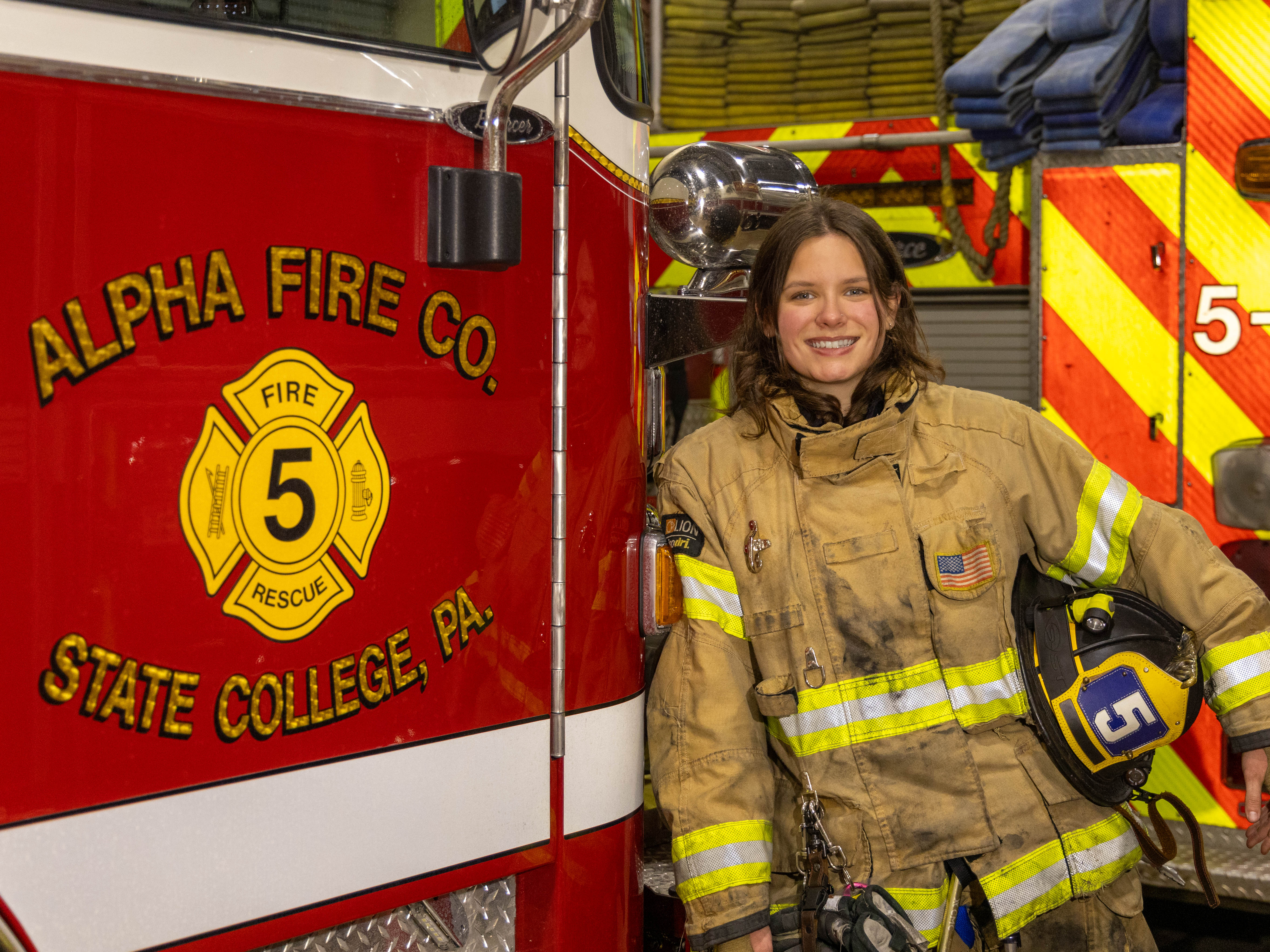 Kara Stover, a Penn State student and volunteer firefighter, leans against a fire engine while wearing her turnout gear.