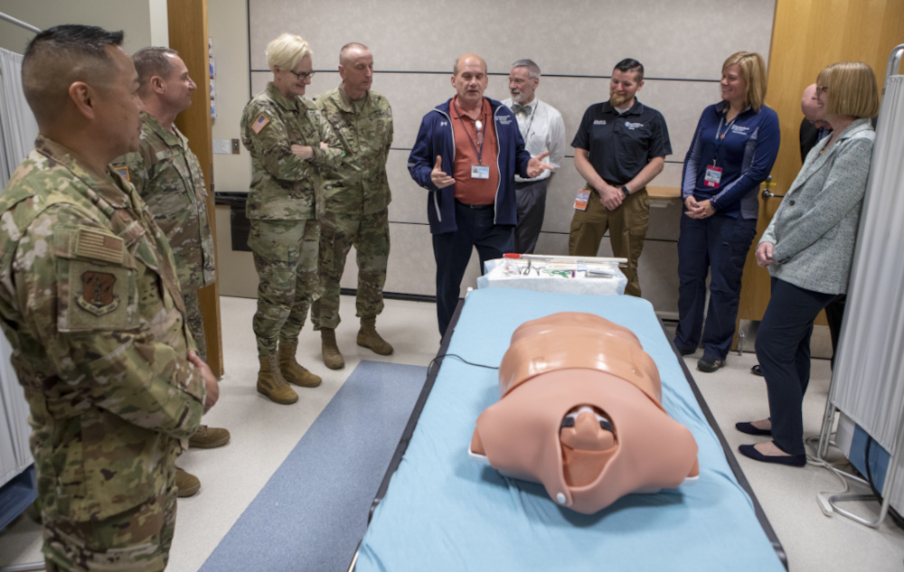 A group of National Guard members in uniform and medical professionals in civilian attire stand around a hospital bed featuring a medical training mannequin. One person in a jacket appears to be explaining or demonstrating something, while the others listen attentively in a clinical setting.