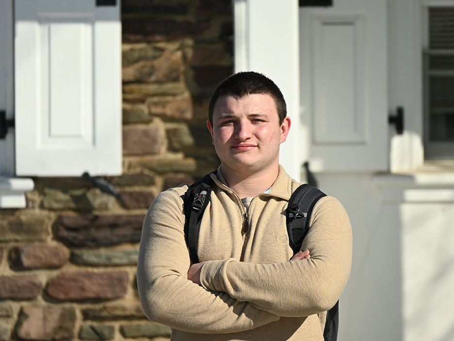 A man wearing a backpacks stands against a stone building with his arms crossed over his chest.