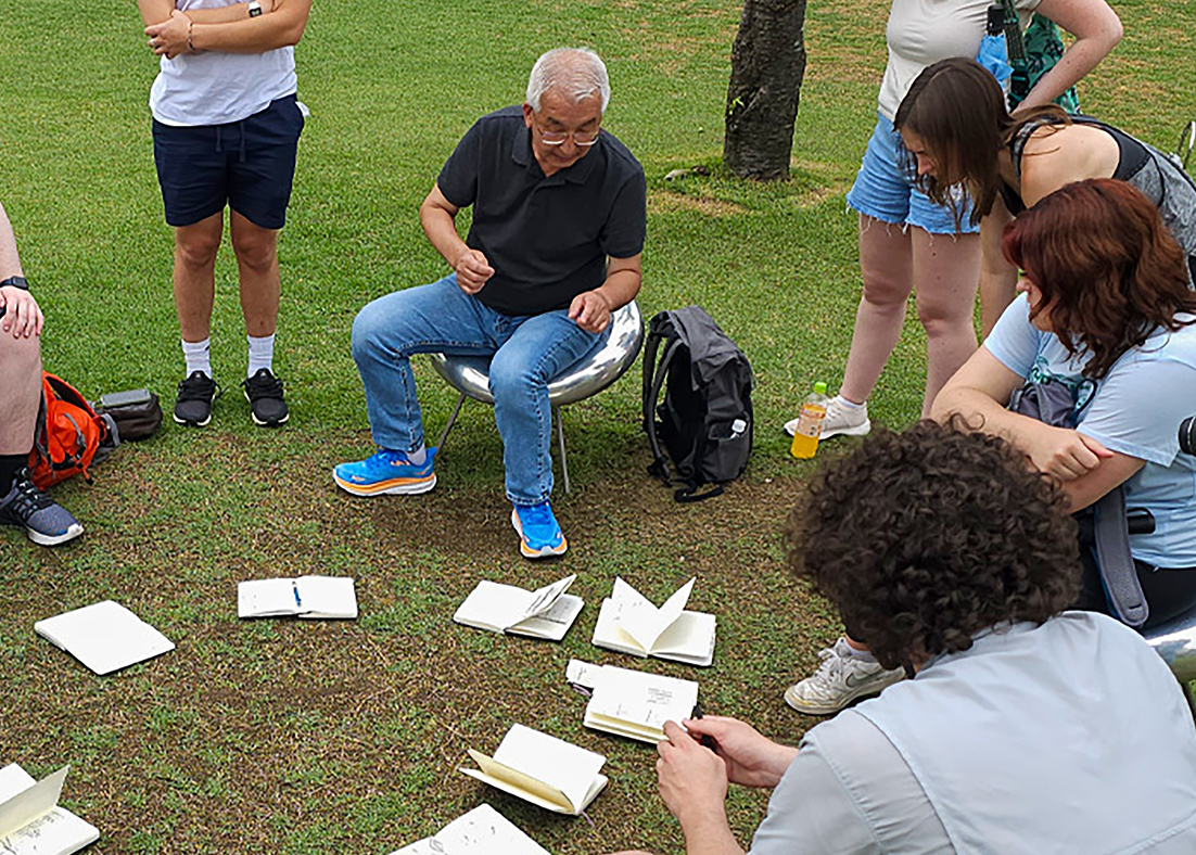 Katsuhiko Muramoto sitting among students outside with sketchbooks open on the ground in front of them.