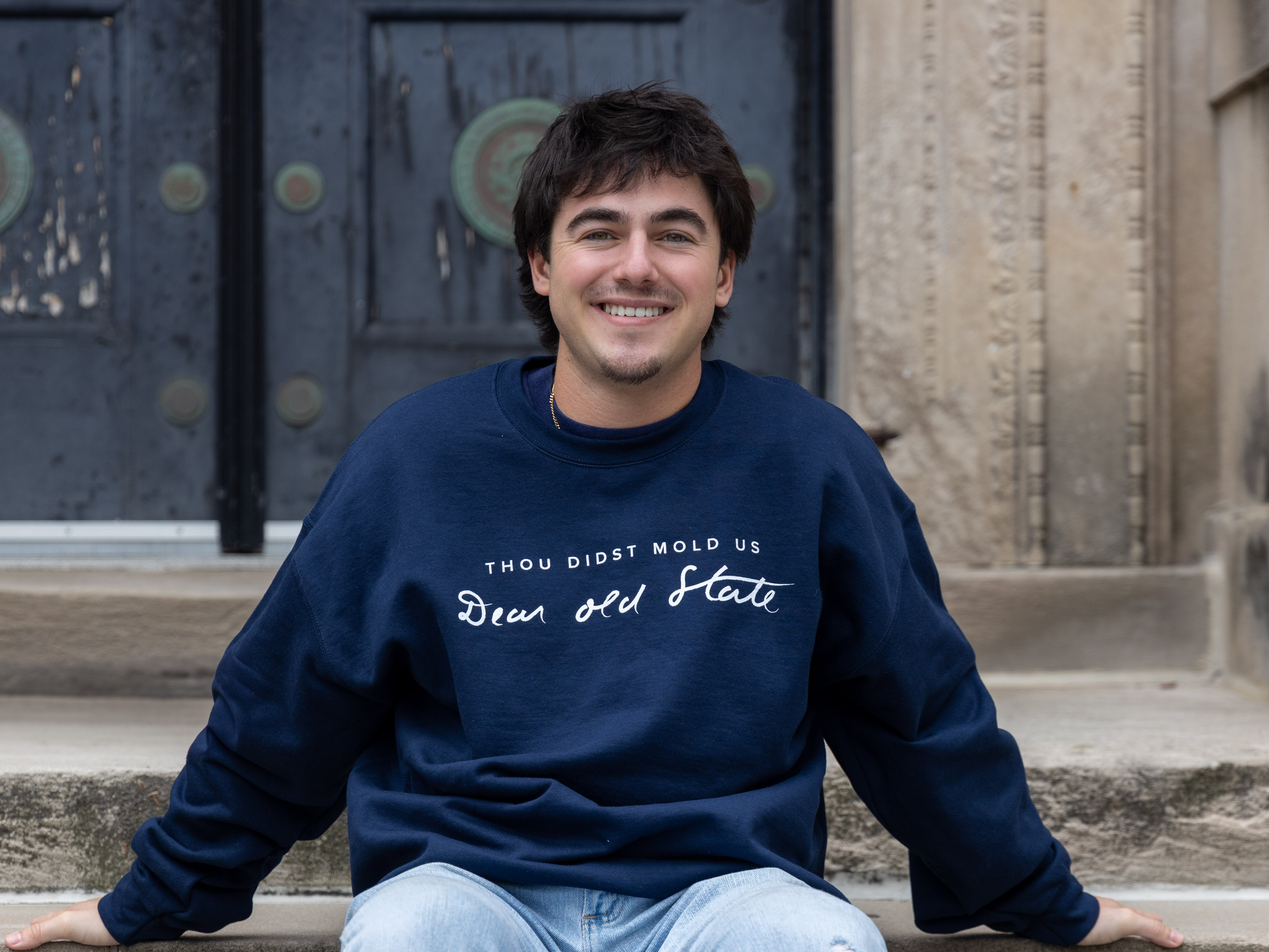 Owen Perry sits on steps outside the main entrance of Sparks Building wearing jeans and a navy sweatshirt that reads “Thou didst mold us Dear Old State.”