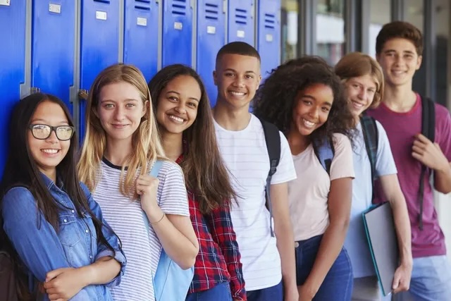 A group of teens stand smiling at the camera against a background of lockers