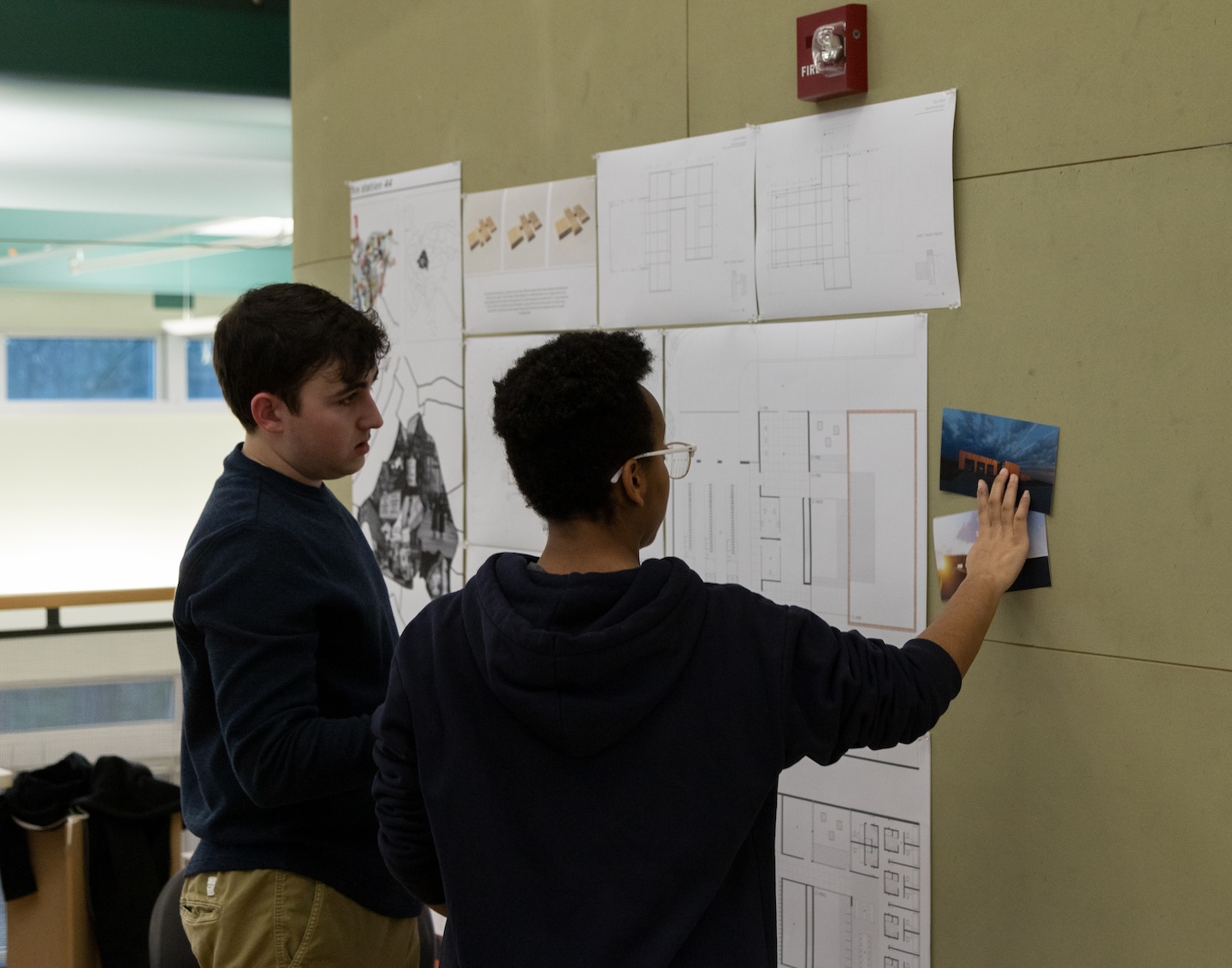 Two students look at architectural designs tacked to a wall.