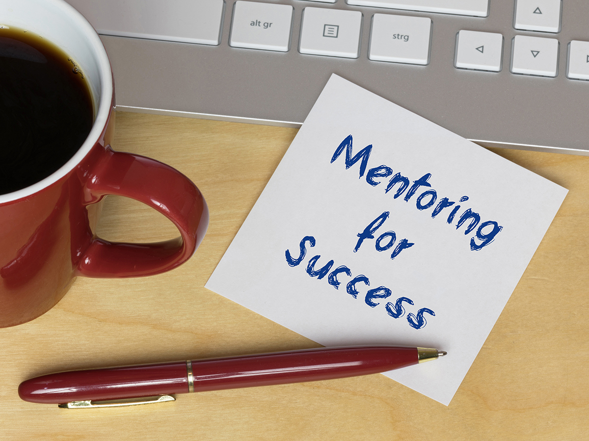 A post-it note reading Mentoring for Success sits on a desk with a pan, a cup of coffee, and a laptop