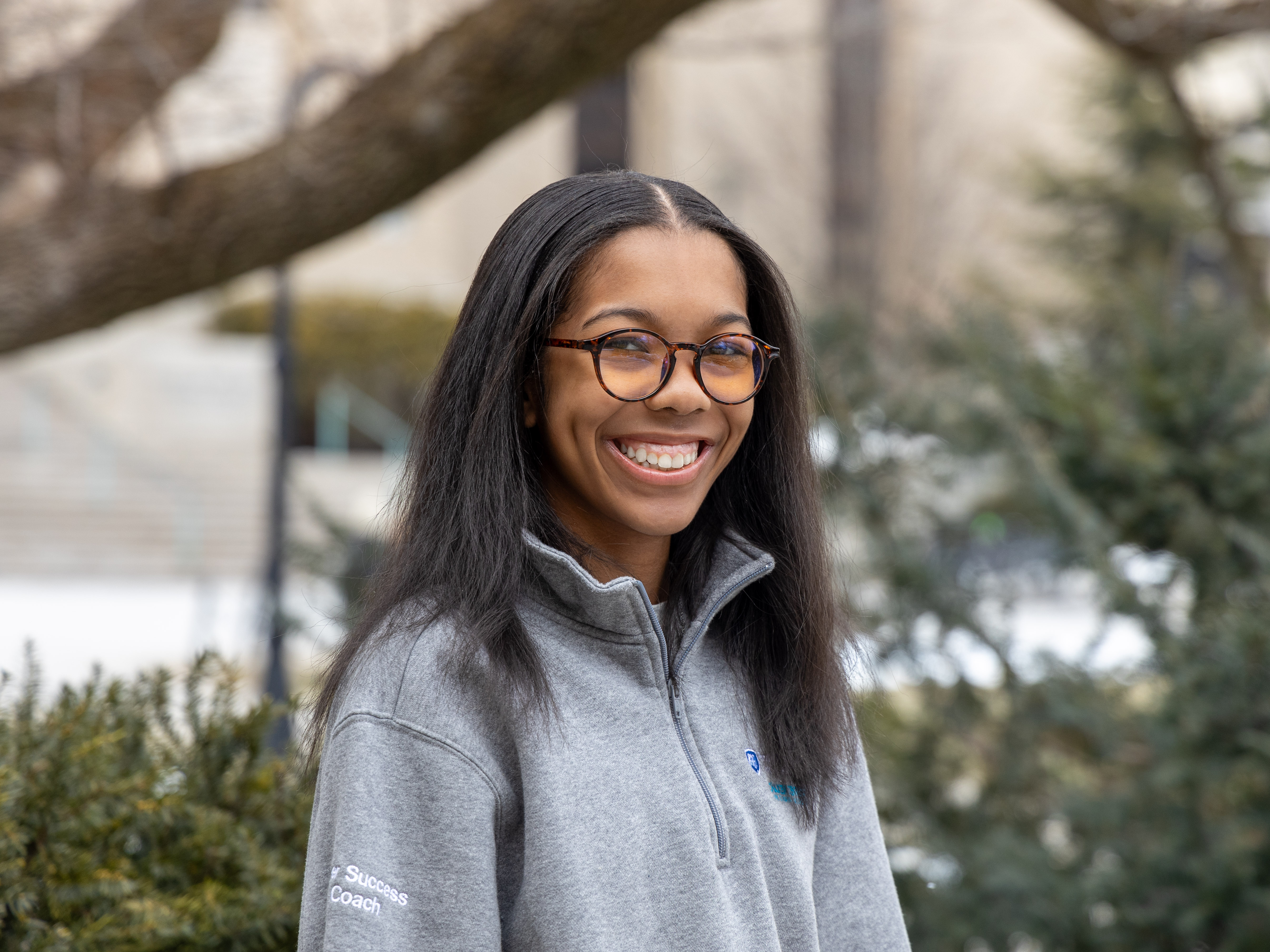 Laci Moore wears a gray Liberal Arts Chaiken Center quarter-zip sweatshirt that says “Peer Success Coach” on the sleeve while standing in front of bushes and trees outside Pattee Library and Paterno Library.