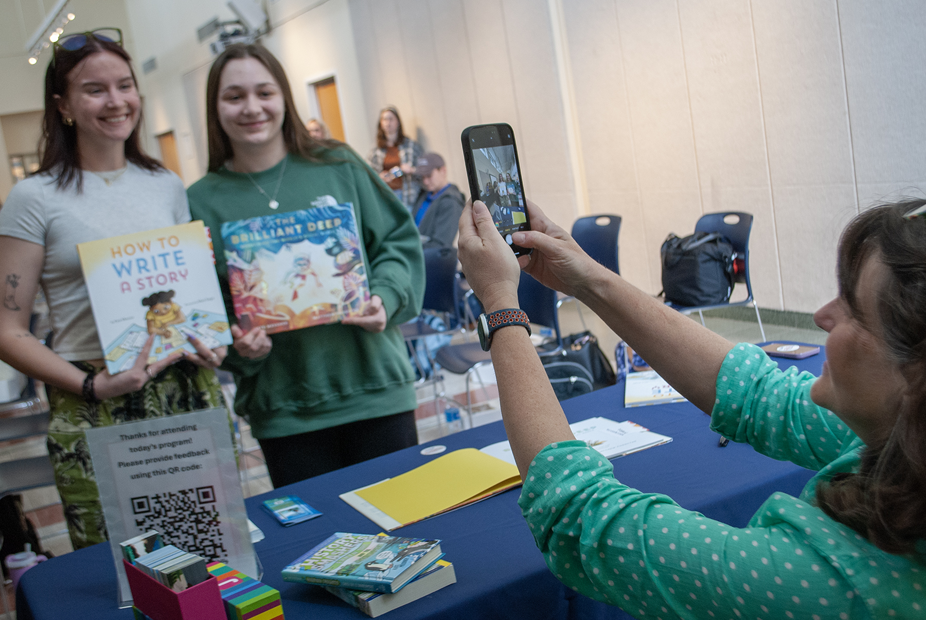 Two students pose holding children's books, while a woman takes a photo with her phone