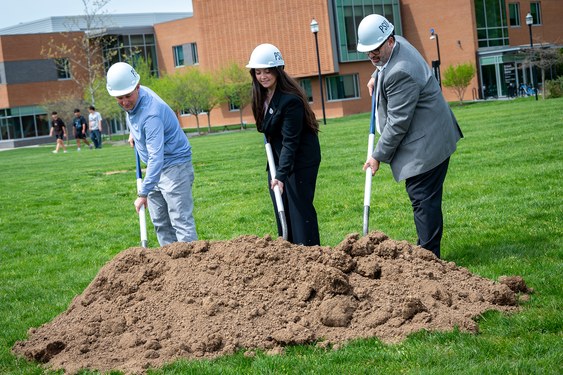 Three people wearing hard hats push shovels into a pile of dirt
