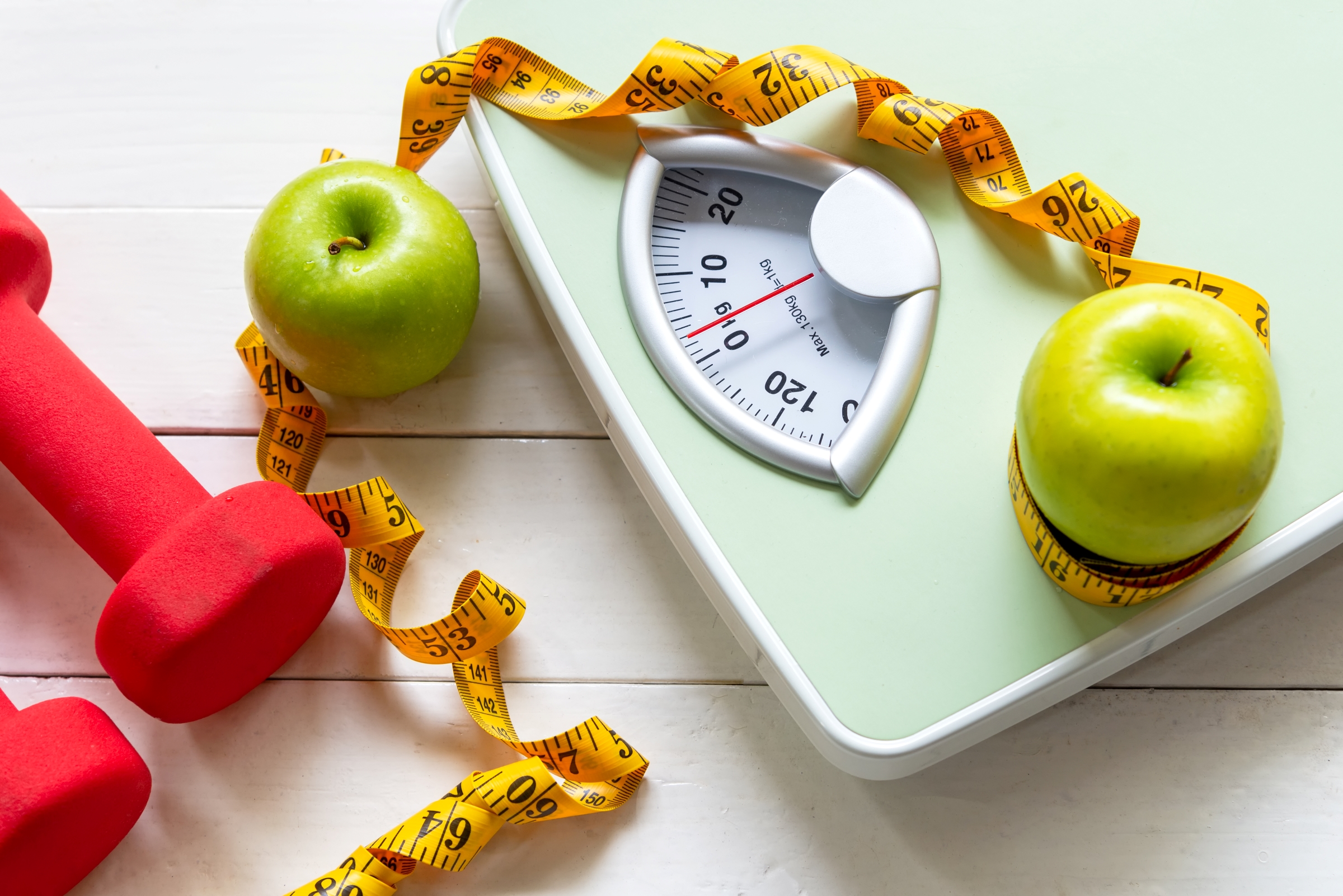 Green colored scale surrounded by two Granny Smith apples, yellow measuring tape next to small red-colored hand weights.