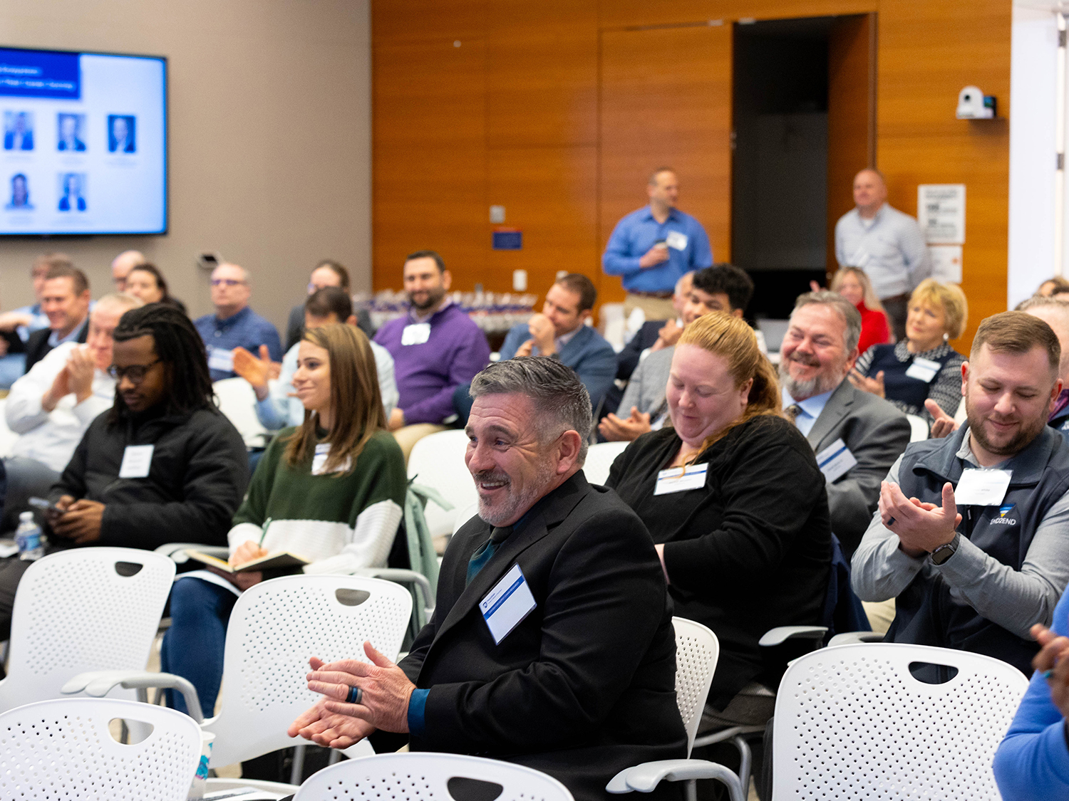 Audience members in a conference room clap and smile.