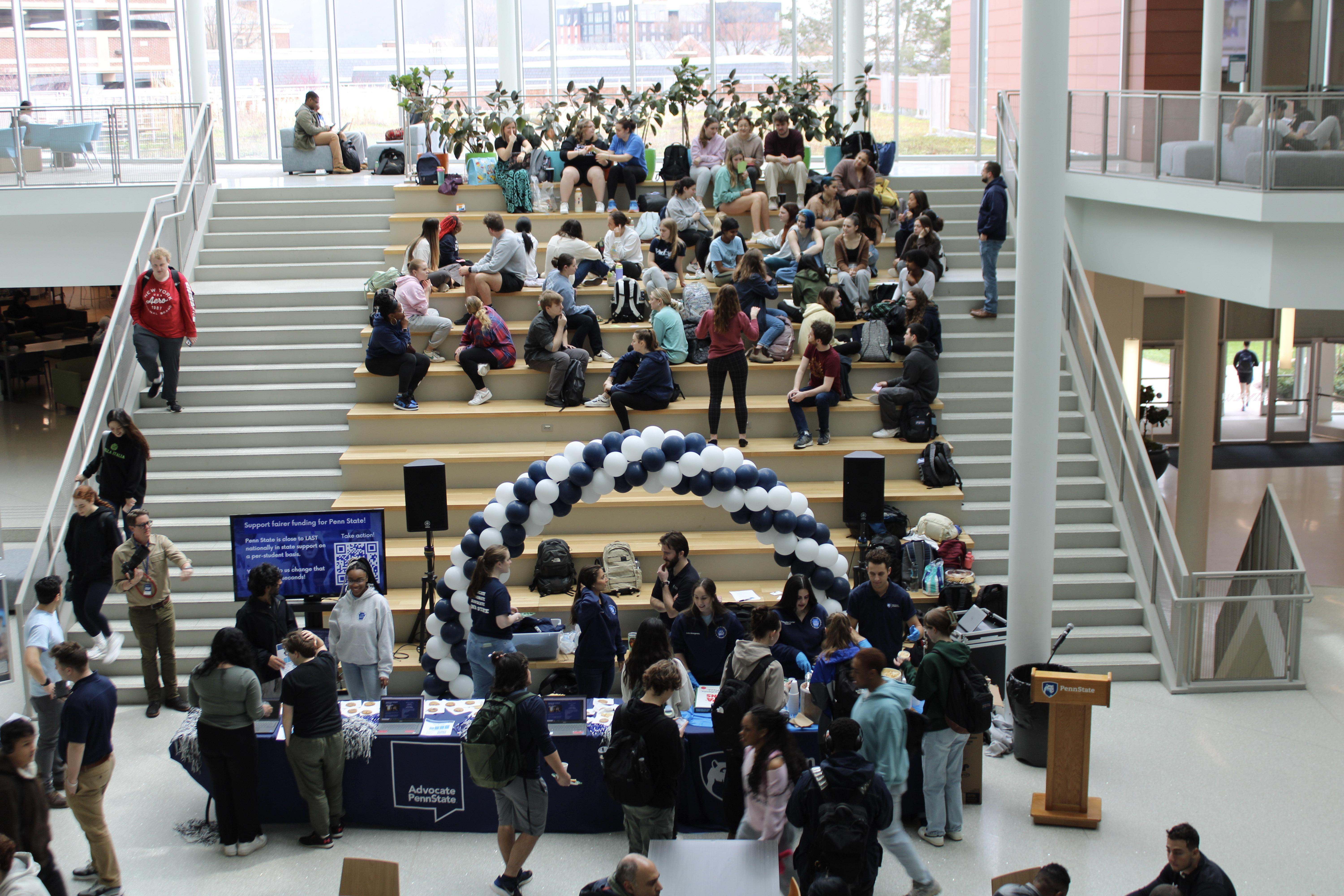 Students gather in a bright, modern atrium with tiered seating for a Penn State advocacy event. A large blue and white balloon arch stands over tables where students interact with organizers, collect materials, and sign up to support fair funding for Penn State. The event features signage, pom-poms, and a podium with the Penn State logo, creating an energetic and welcoming atmosphere for civic engagement