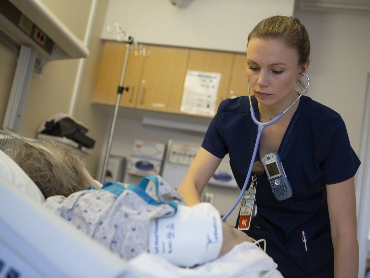 A nurse uses a stethoscope as she tends to a young patient in a hospital bed. The nurse also has a phone and a nametag clipped to her shirt. An IV pole and wooden cabinets are in the background.