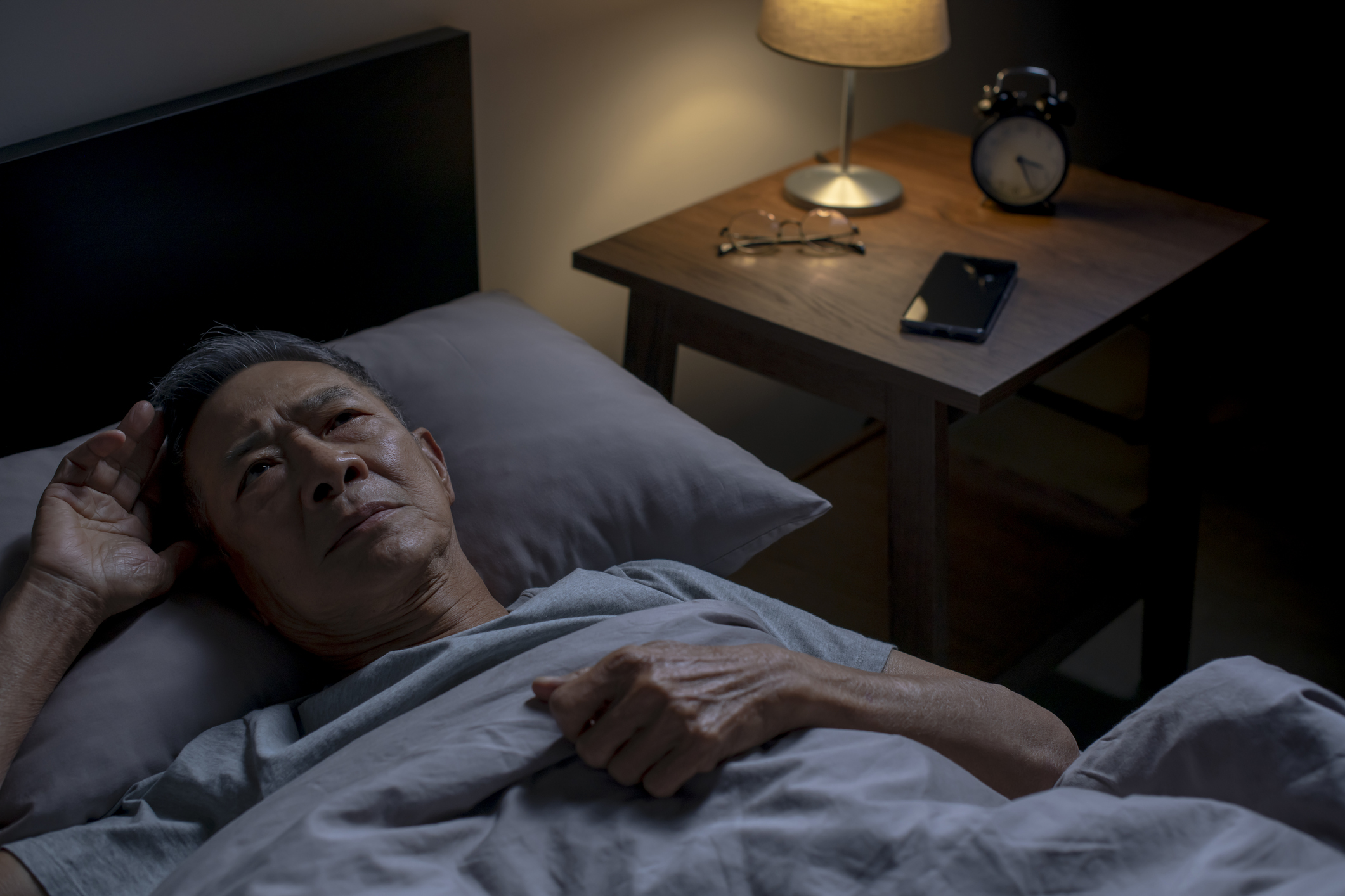 An older person in bed in a dark room stares at the ceiling, looking uncomfortable. A small bedside light is on, and the clock reads 3:25. 
