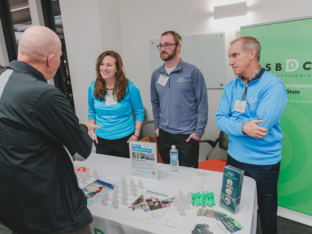 Three individuals stand behind a table with SBDC materials, speaking to an individual on other side of table.