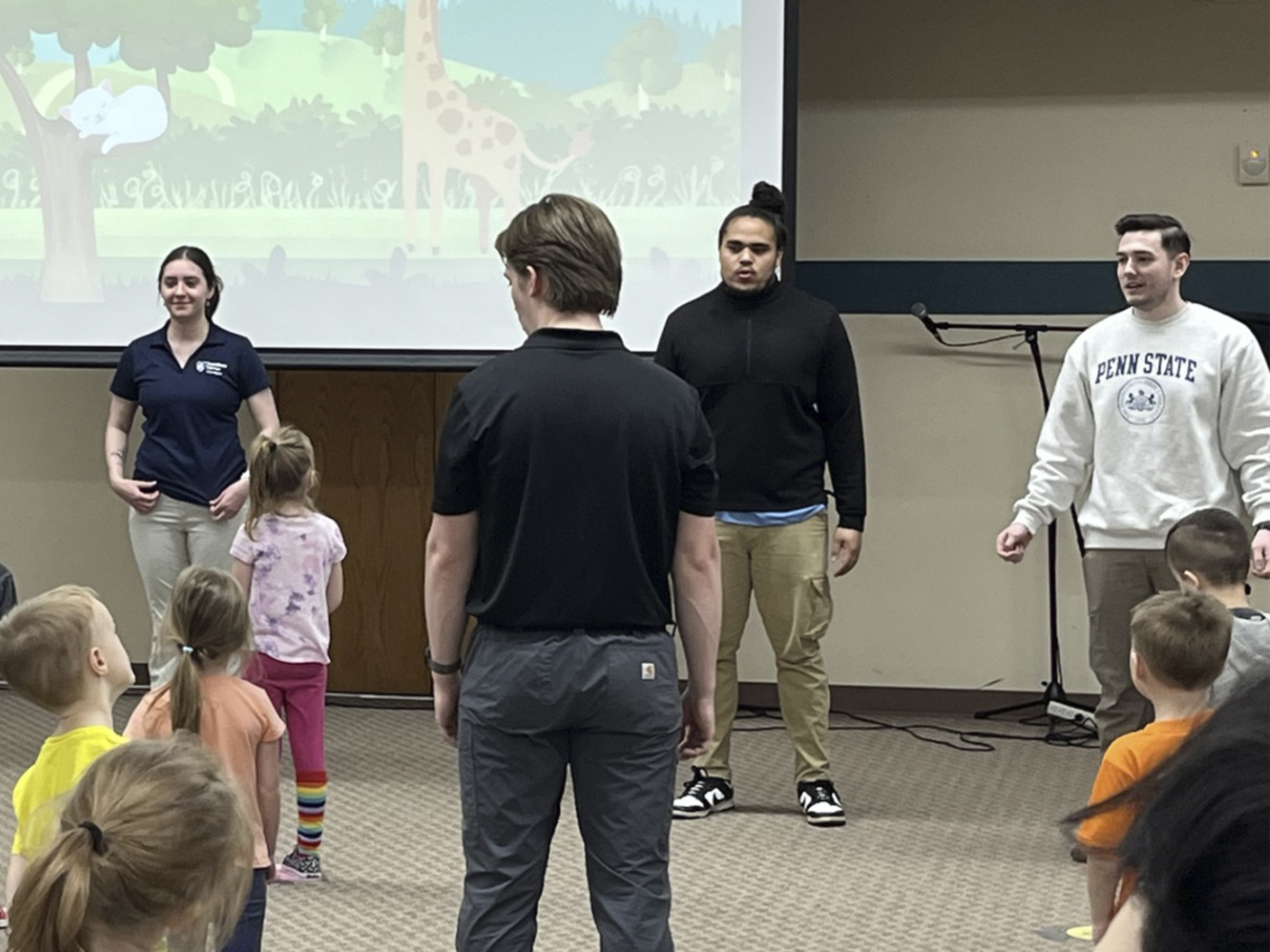 Students standing in front of a projector screen speaking demonstrating for preschool children