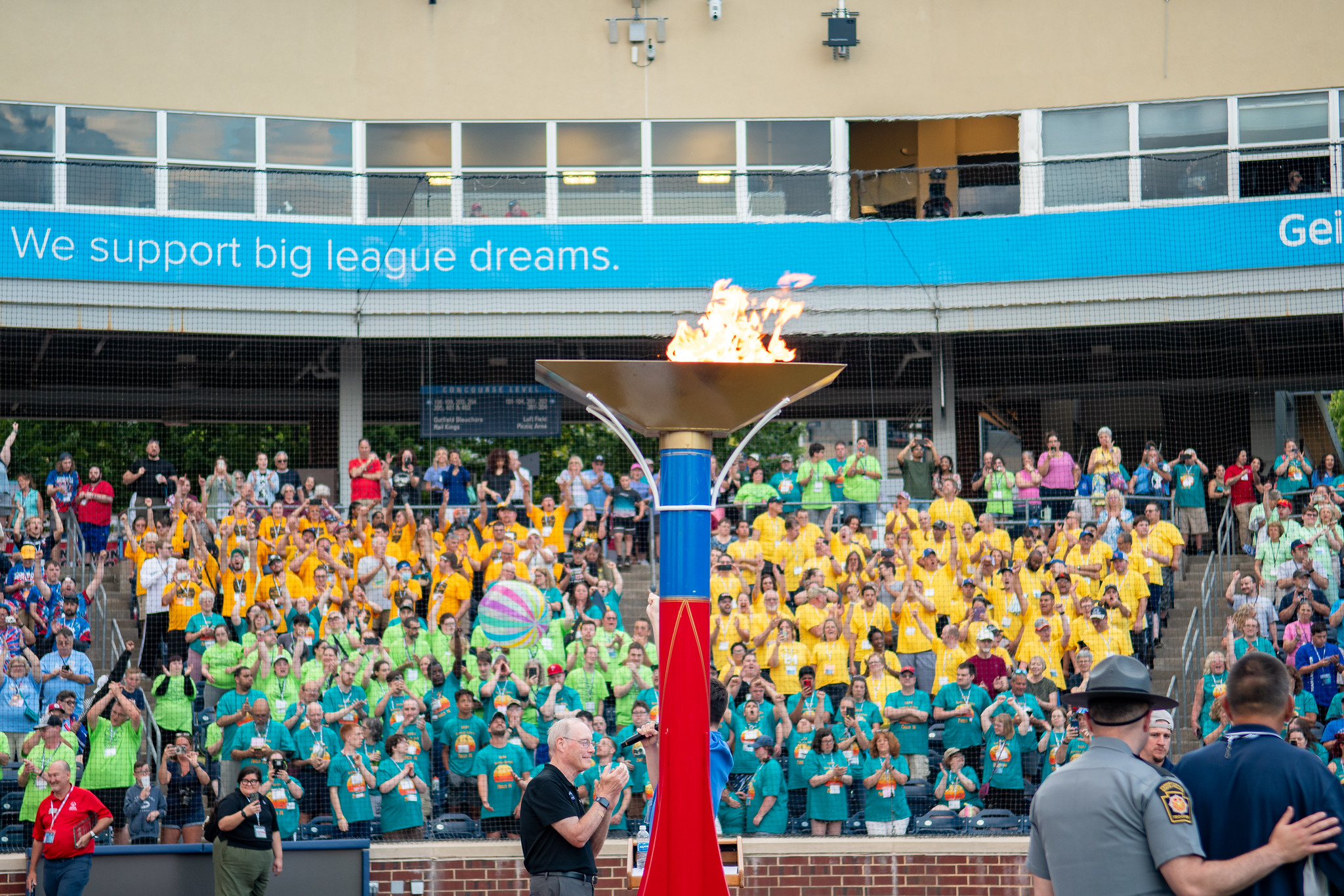 A huge lit torch is backgrounded by stadium stands full of people wearing colorful t-shirts