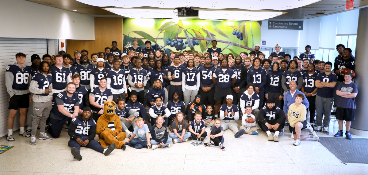 A large group photo of the Penn State football team, all wearing navy blue jerseys, posing with children and staff in a brightly lit hospital lobby.