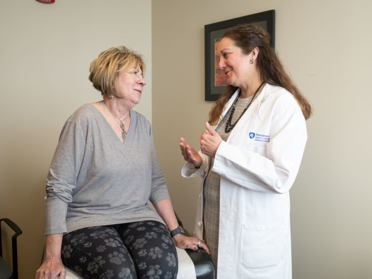 A woman sits on an exam table talking to Jaime Long, right, of Penn State Health. Long is gesturing with her hands and wearing a white coat with the Penn State Health Milton S. Hershey Medical Center logo on it. Both are smiling.