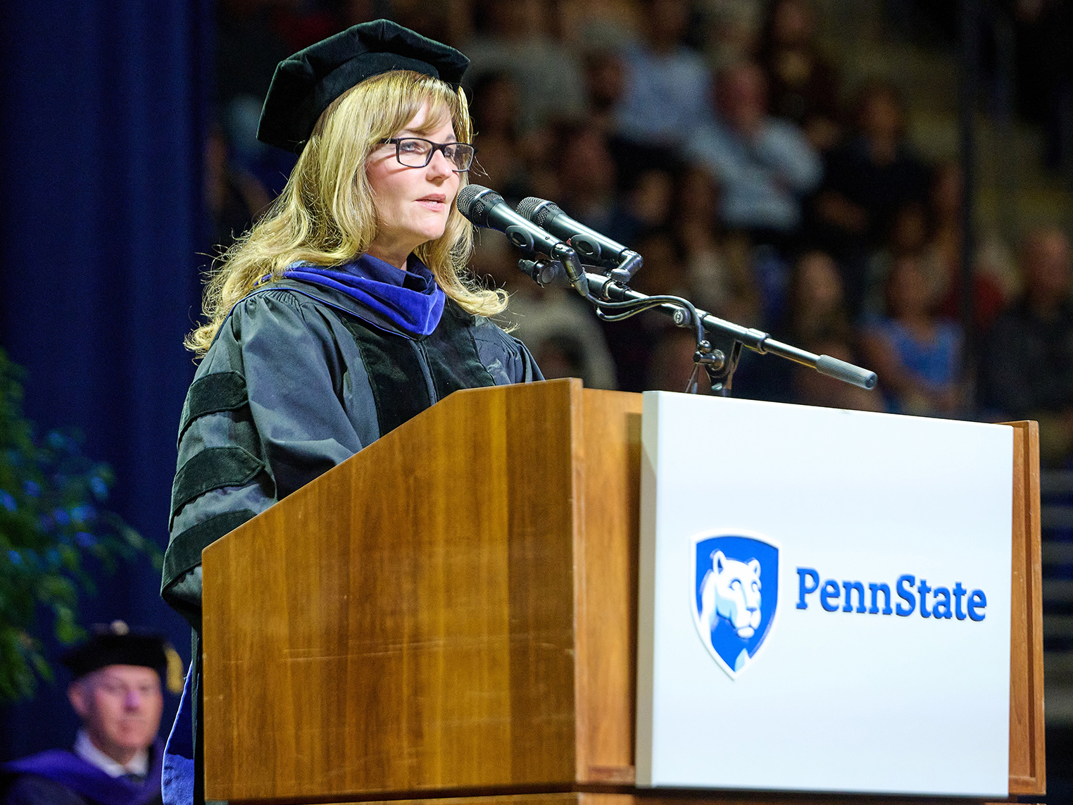 A photo of DuPont CEO Lori Koch in a cap and gown speaking behind a podium with a Penn State graphic during Smeal's spring 2025 commencement ceremonies.