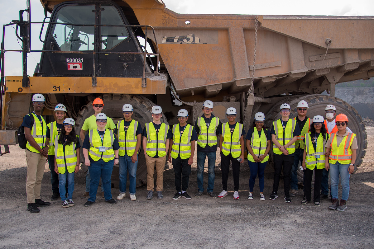 A group of students wearing safety helmets and high-visibility vests stand in front of a large yellow industrial truck at quarry.
