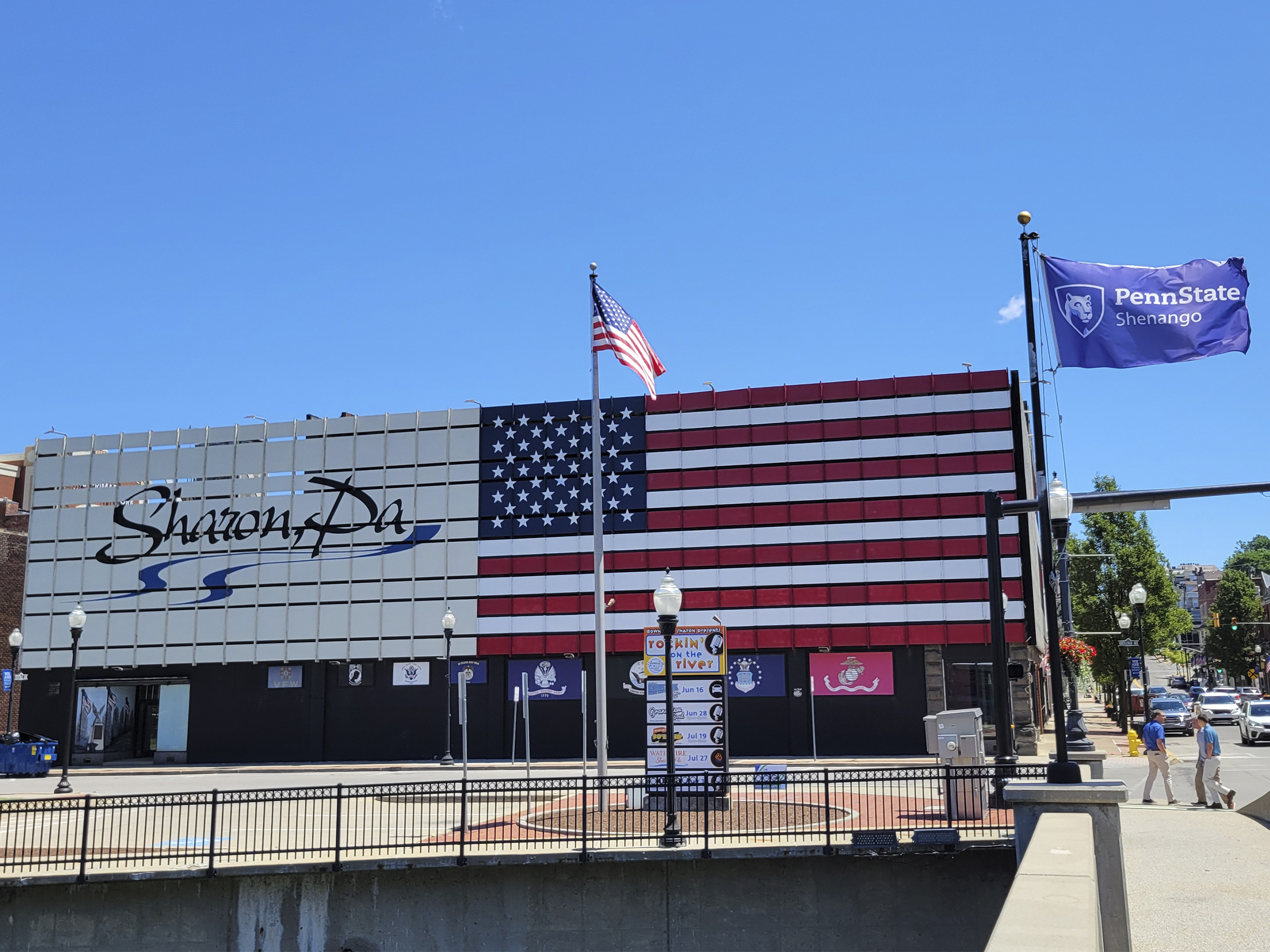 Exterior of a building with an American flag painted on the side and text saying Sharon, PA with sidewalks and flag poles in the foreground