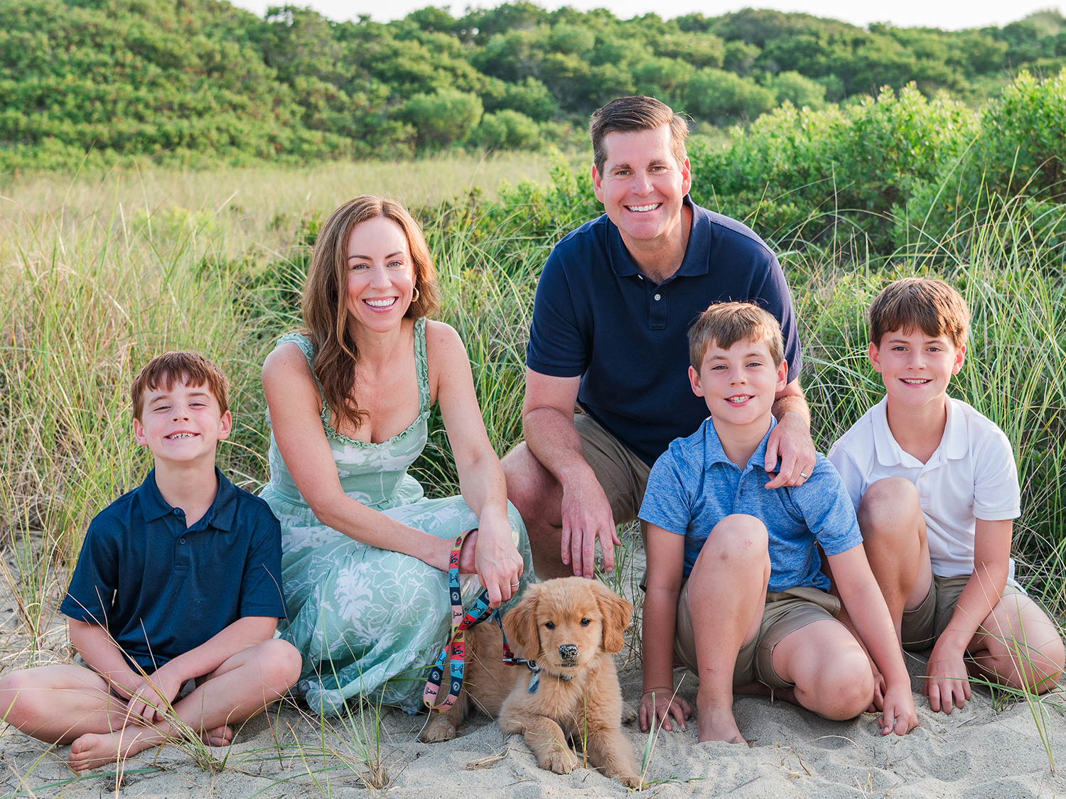A photo of the Brandau family posing in sand in front of ocean grass. Jim and Micaela are shown with young sons Jack, Charlie and Max.